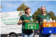 Zwei unserer Fahrer*innen tragen gepackte Kisten in grün (Obst und Gemüse), orange (Trockenprodukte) und blau (Kühlprodukte)