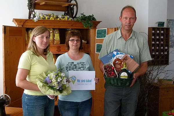 2 Frauen und ein Mann mit Blumenstrauß, Karte von Regionales Bayern und Geschenkkorb in den Händen.